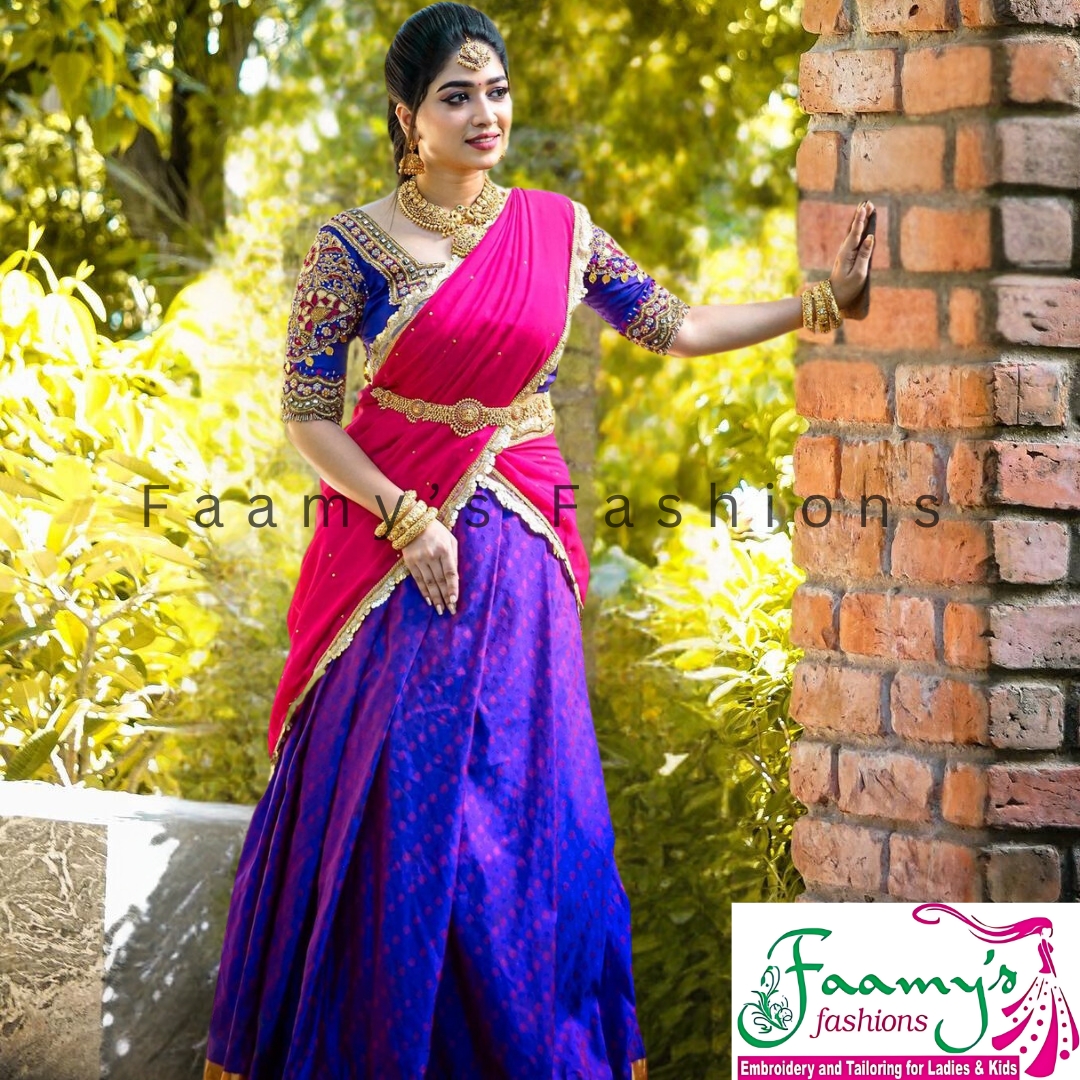 Elegant woman in vibrant pink and blue traditional Indian saree with intricate embroidery, posing beside a brick wall amidst lush greenery.