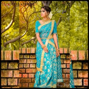 Elegant woman in a turquoise blue saree with golden embroidery, standing by a brick wall in a garden.